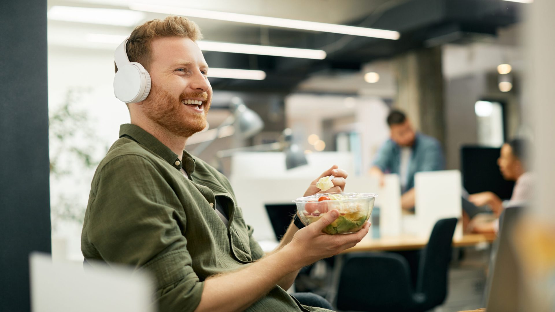 Happy entrepreneur enjoying while eating lunch and listening music over headphones in the office.